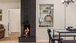 A modern living room with a black brick fireplace, a dining table with chairs, a chandelier, and a glass display cabinet with decorative items. - Beach St Escape-Hot tub-5min Walk to the Beach (Muskegon)
