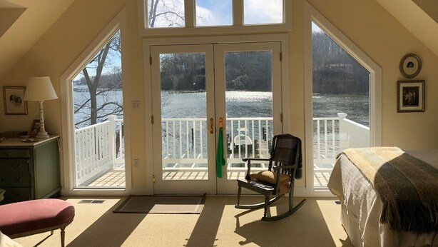 Ein Zimmer mit großem Erkerfenster, das auf einen See blickt, einem Schaukelstuhl und einem Bett.