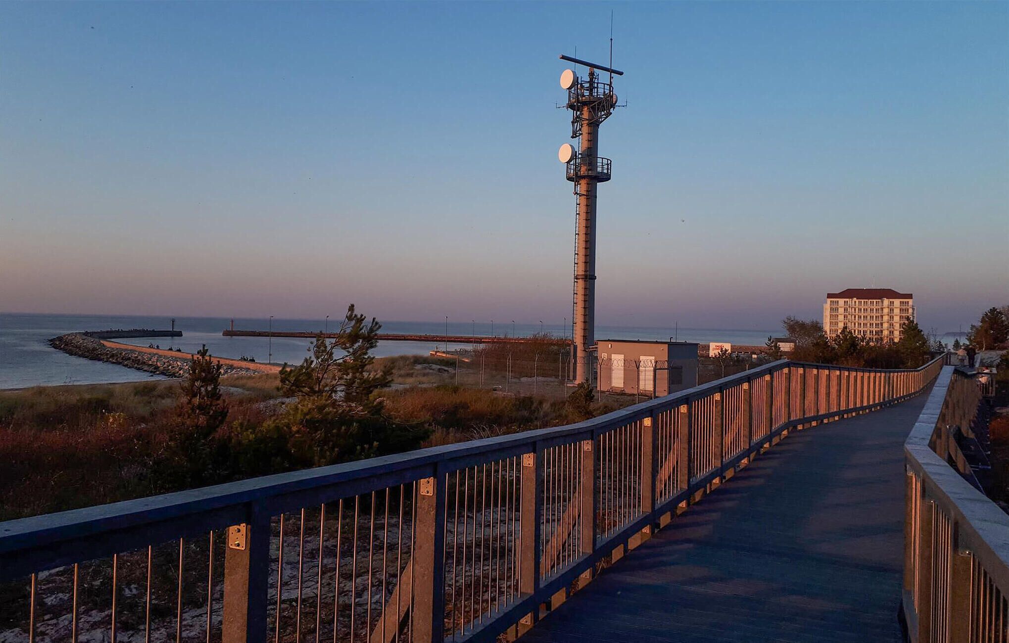A coastal walkway with a metal fence, a tall communication tower, and a building with a red roof in the background.