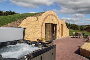 A hot tub area with a stone building, outdoor seating, and a grassy hill in the background.