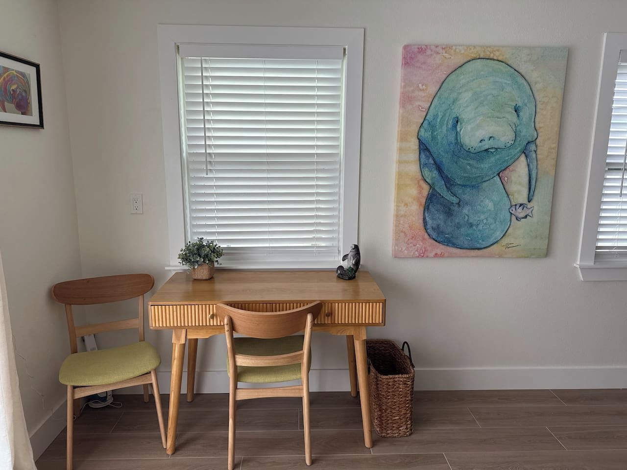 Una habitación con un escritorio de madera, dos sillas, una ventana con persianas, un cuadro colgado en la pared y una cesta de mimbre.