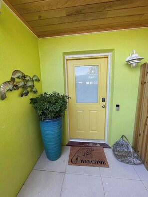 A yellow door with a welcome mat, a potted plant, and a wall-mounted light fixture.