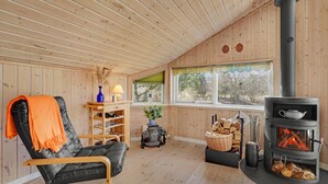 A cozy cabin interior with a wooden interior, a black leather chair with an orange throw, a wooden shelf with books and a vase, a window with a view of trees, and a wood-burning stove.
