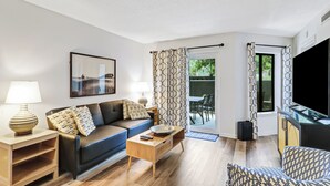 A living room with a sofa, coffee table, and a TV. There is a door leading to an outdoor area with a table and chairs. - Walk to beach, updated 2/2 condo sleeps 6, in & outdoor pools, tennis, courtyard (Hilton Head Island)
