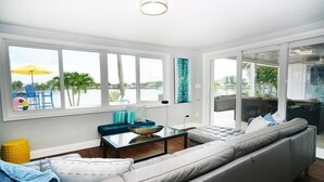 A modern living room with a large gray sofa, a glass coffee table, and a view of the water. - Fabulous views, heated pool 5 BR 5.5 BA Waterfront (Indian Rocks Beach)