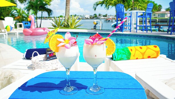 Two cocktails on a blue table by a pool with lounge chairs and umbrellas. - Fabulous views, heated pool 5 BR 5.5 BA Waterfront (Indian Rocks Beach)