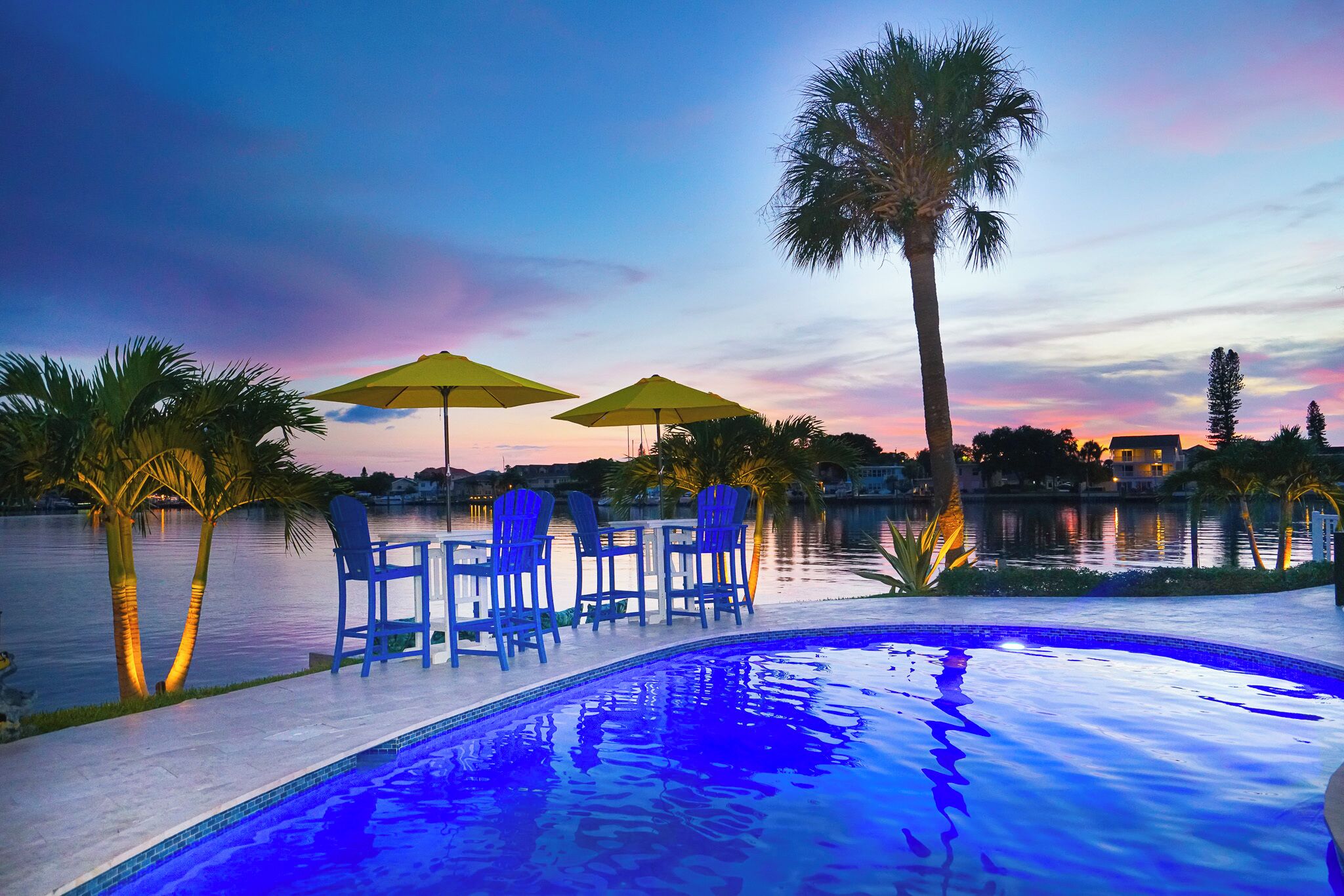Zona junto a la piscina con sillas y sombrillas, una palmera y vista a una masa de agua al atardecer.