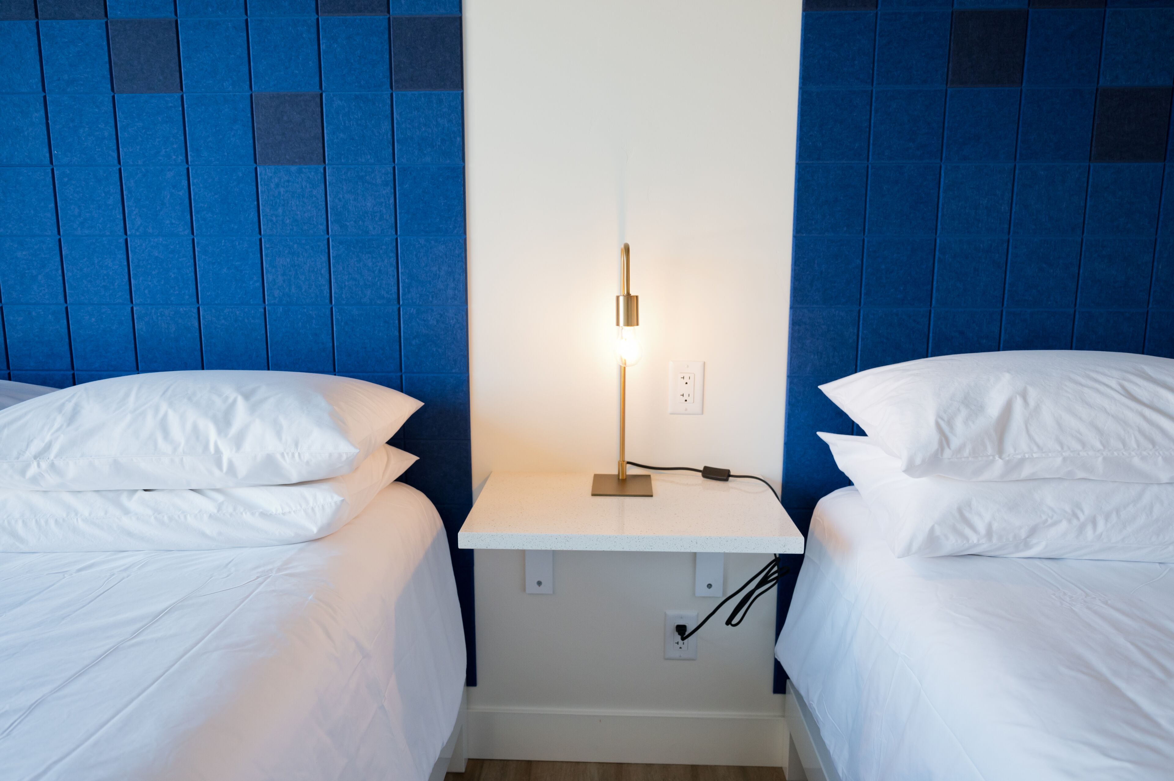 A hotel room with two beds, a nightstand with a lamp, and a wall with blue tiles.
