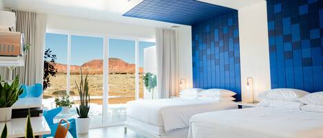 Habitación de hotel moderna con dos camas, zona de comedor con mesa y sillas, y un ventanal con vistas al desierto.