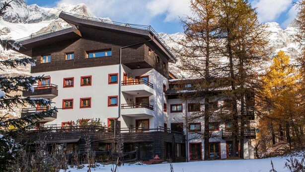 A multi-story hotel with a modern design, featuring balconies and large windows, set against a backdrop of snow-covered mountains and autumn-colored trees. - Residence Cervinia ID 522 (Valtournenche)