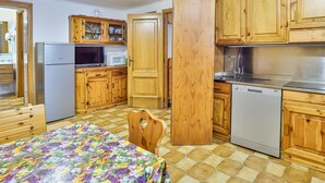 A compact kitchen with wooden cabinets, a refrigerator, a microwave, a dishwasher, and a dining table with a floral tablecloth. - Residence Cervinia ID 522 (Valtournenche)