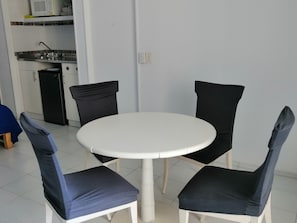 A small dining area with a white round table and four chairs, each with a black cushion. A kitchen area with a microwave and sink is visible in the background.