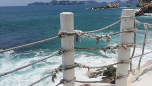 Vista costera con una barandilla de cuerda y un pilar blanco, con vista al mar y la tierra distante.