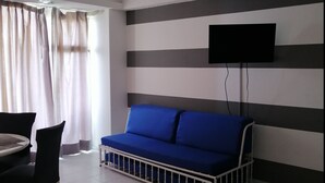 Una sala moderna con un sofá azul, un televisor de pantalla plana montado en la pared, un ventilador de techo y un comedor con mesa y sillas.