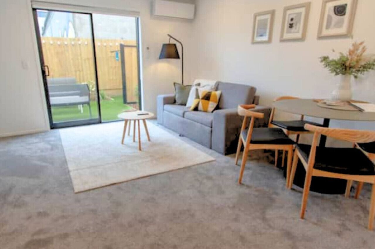 Ein Wohnzimmer mit grauem Sofa, einem kleinen runden Tisch und zwei Holzstühlen. Eine Glastür führt auf eine Terrasse.