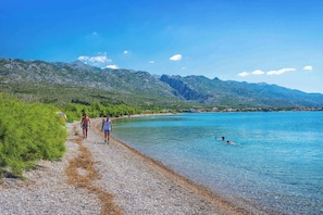 A pebbled beach with clear water, mountains in the background, and a few people enjoying the scenery.
