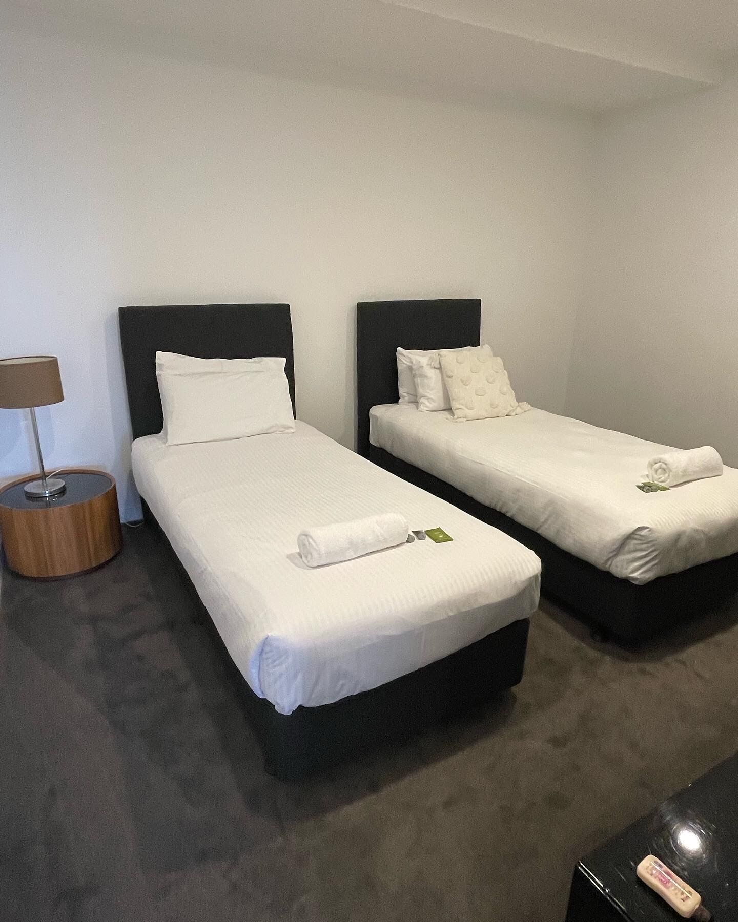 Camera d'albergo con due letti singoli, ognuno con copripiumino e cuscini bianchi, un comodino con una lampada e un tavolino rotondo con una bottiglia.