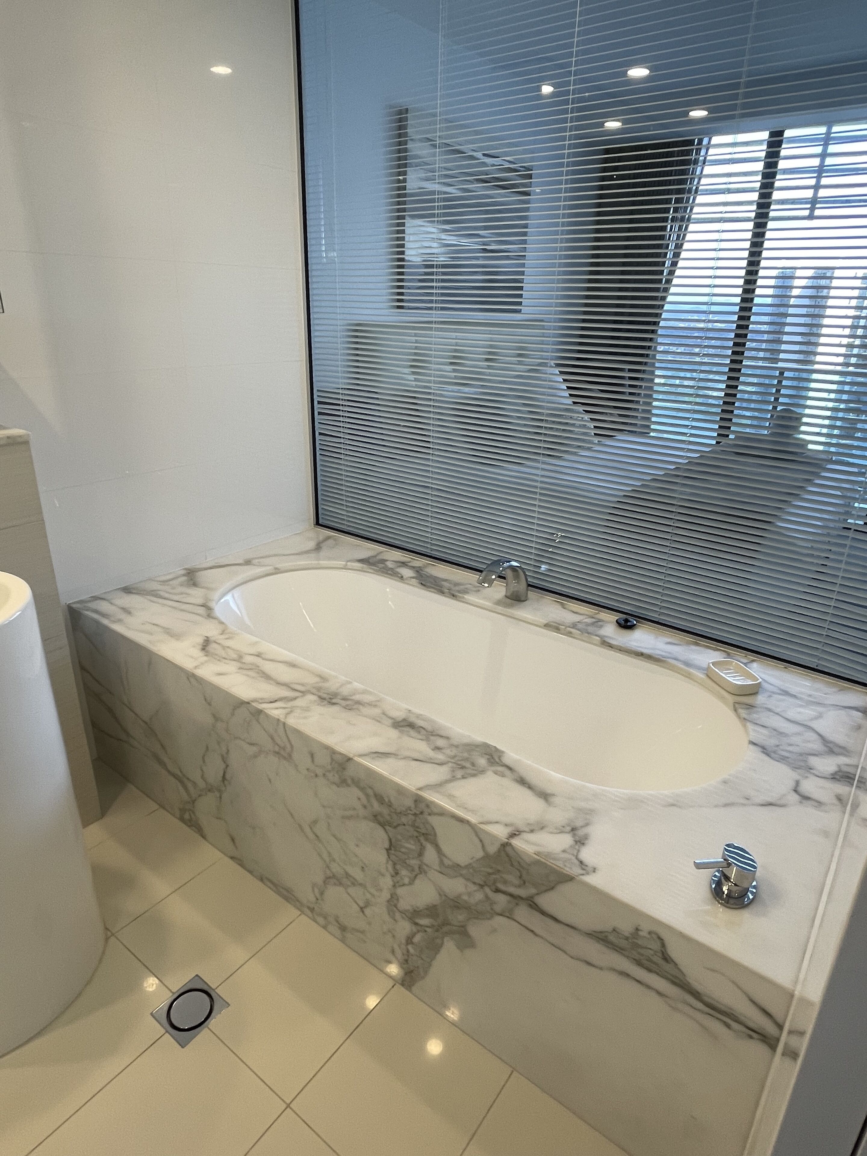 Ein modernes Badezimmer mit einem Marmorwaschtisch, einer freistehenden Badewanne und einem großen Fenster mit Jalousien.