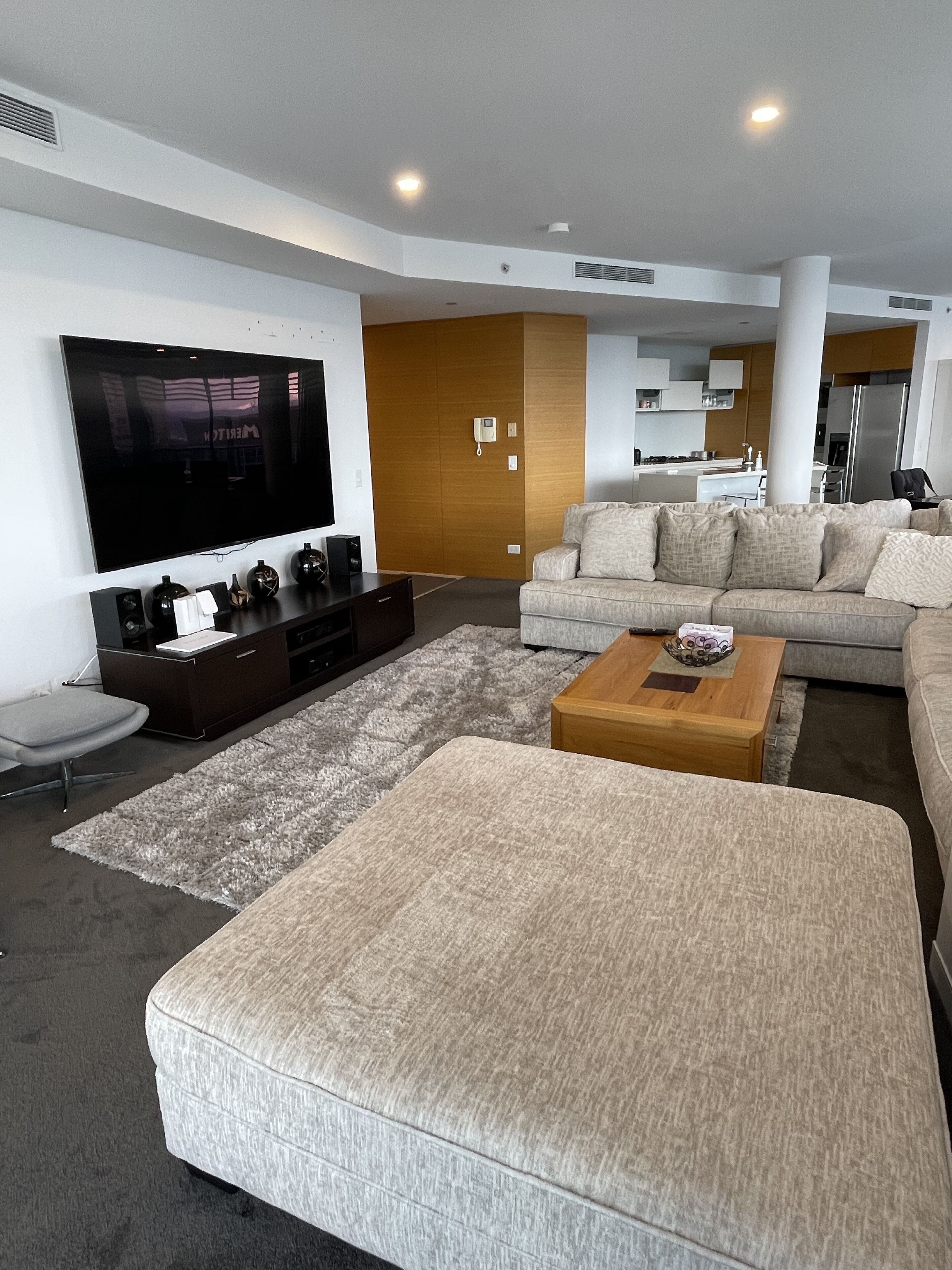 A modern living room with a large flat-screen TV, a sofa, a coffee table, and a kitchen area in the background.