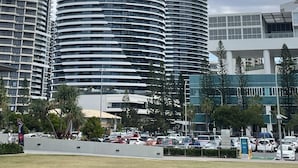 Um arranha-céu moderno e curvo, com várias varandas e fachada de vidro, cercado por outros edifícios e um estacionamento.