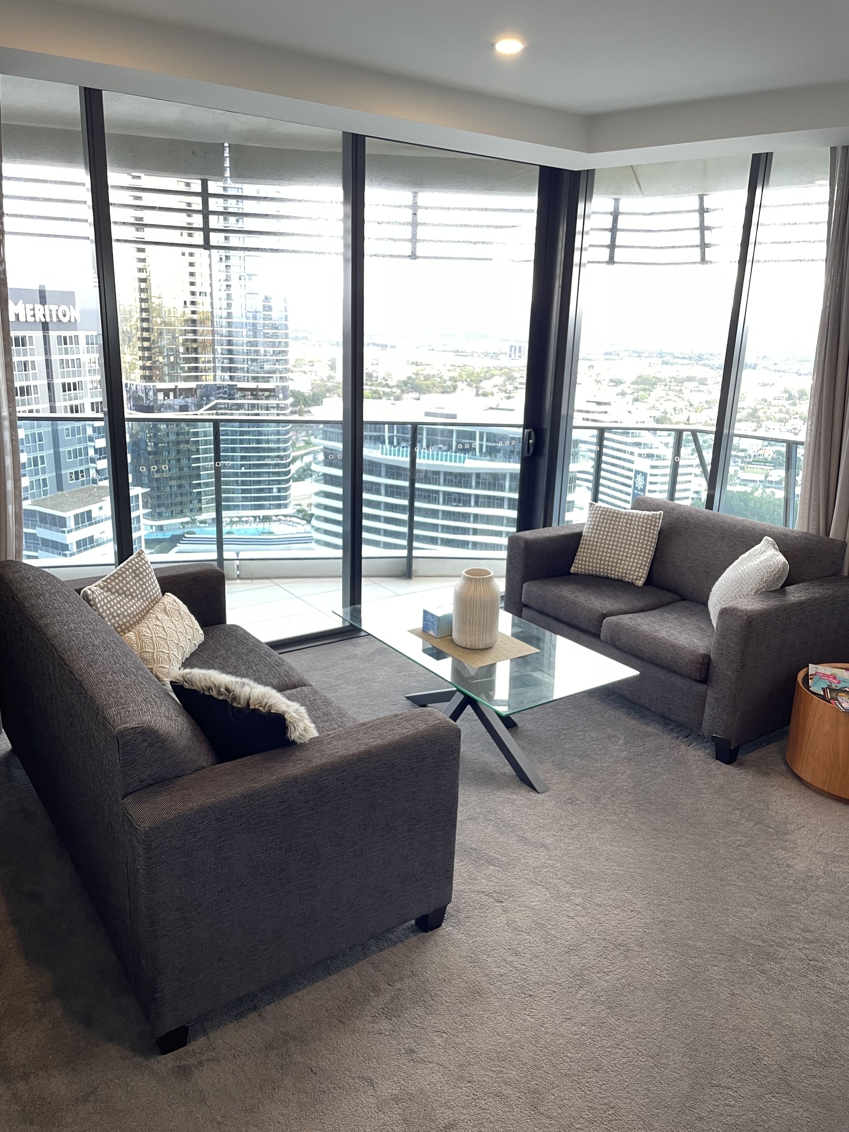 A modern living room with two grey sofas, a glass coffee table, and a view of a cityscape through large windows.