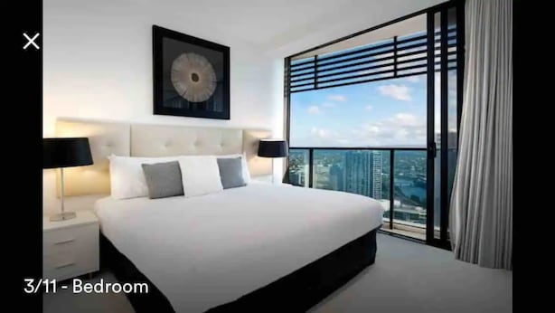 Quarto com uma cama grande, cabeceira, mesa de cabeceira, relógio na parede e vista para a cidade através de janelas do chão ao teto.