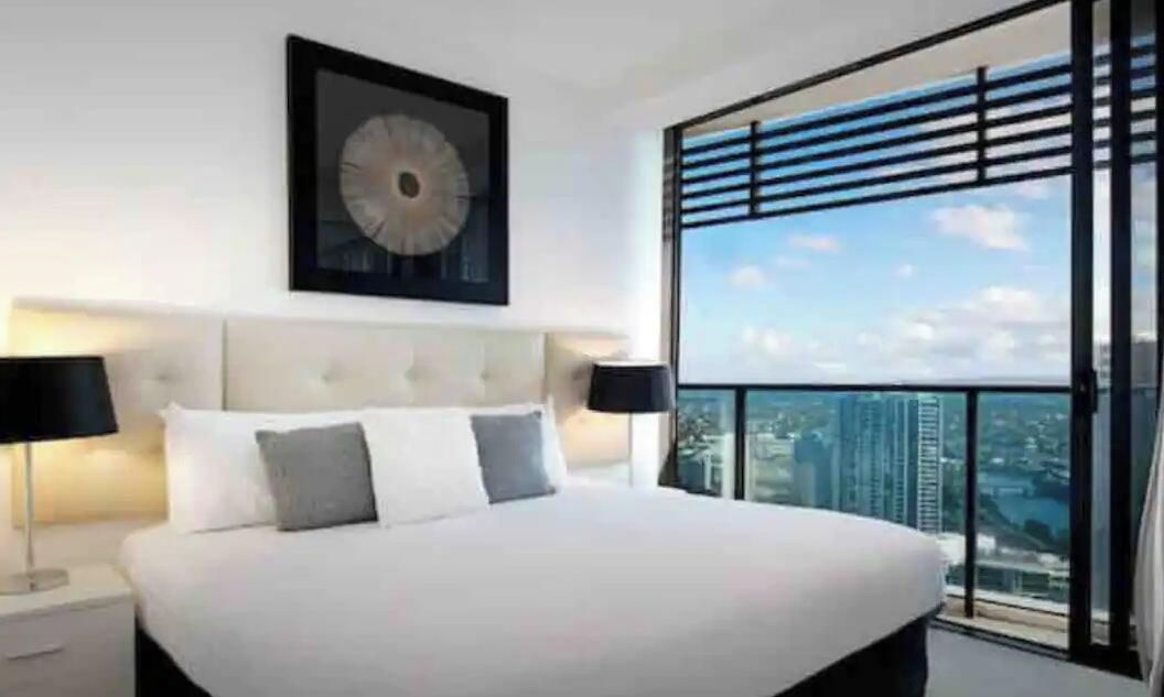 A bedroom with a large bed, a headboard, a nightstand, a wall-mounted clock, and a view of the city through floor-to-ceiling windows.