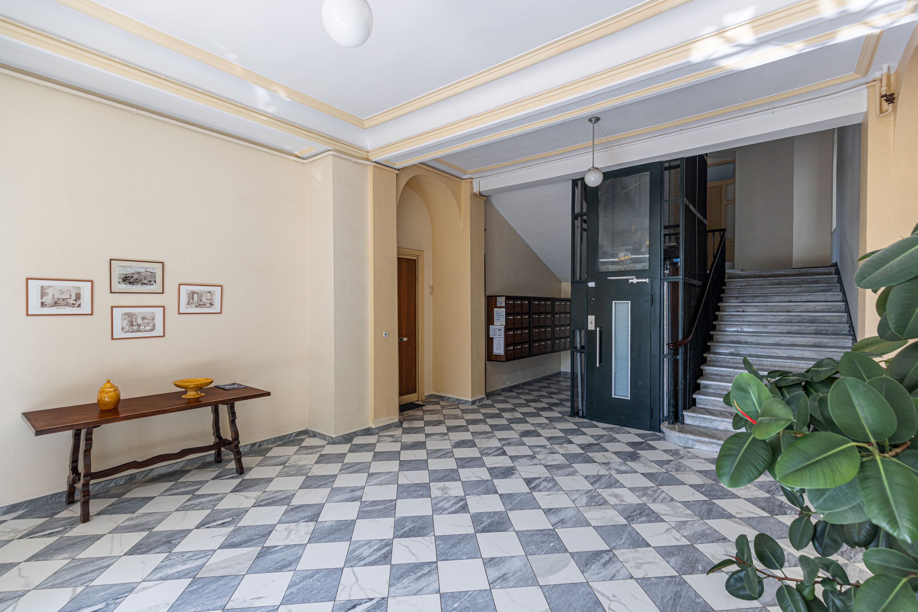 A hallway with a checkered floor, a wooden bench, framed pictures on the wall, and a staircase.