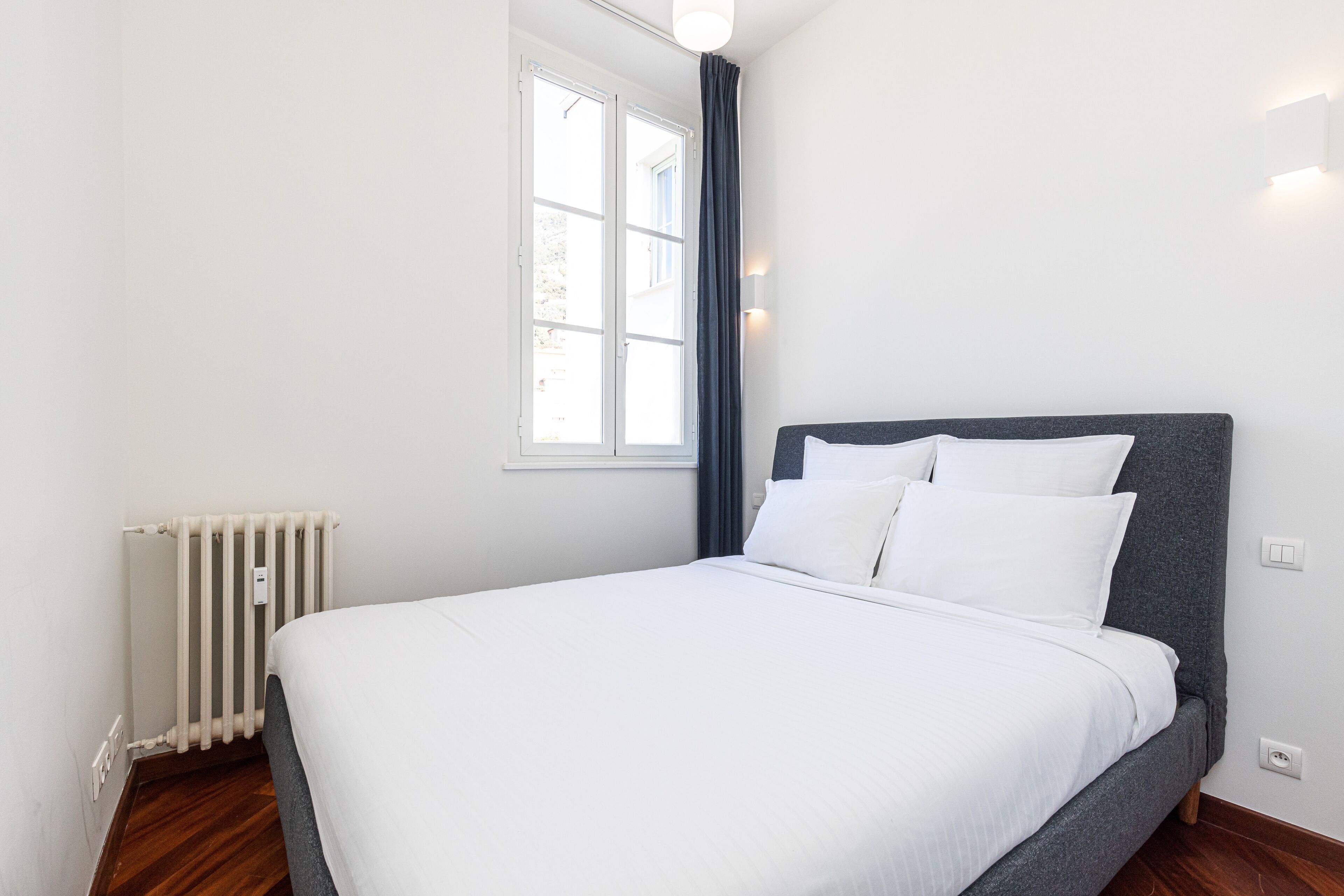 A bedroom with a large bed, a window with curtains, a radiator, and a wall-mounted light fixture.