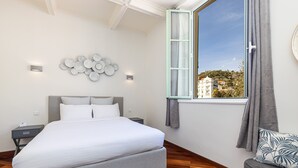 A bedroom with a large bed, a nightstand, a sofa, a window with a view of buildings, and wall-mounted lights. - Splendid sea view apartment (Menton)