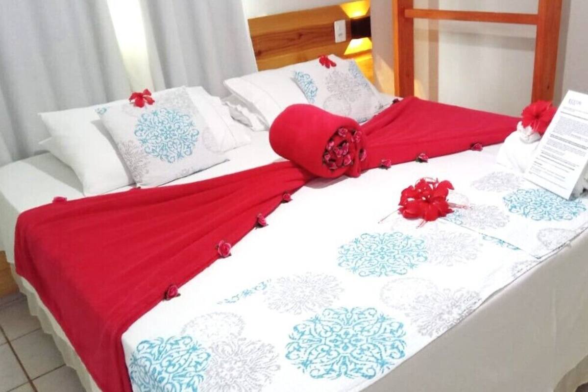 Una cama bien hecha con una colcha roja y almohadas, un cabecero de madera y una pared blanca con una ventana.