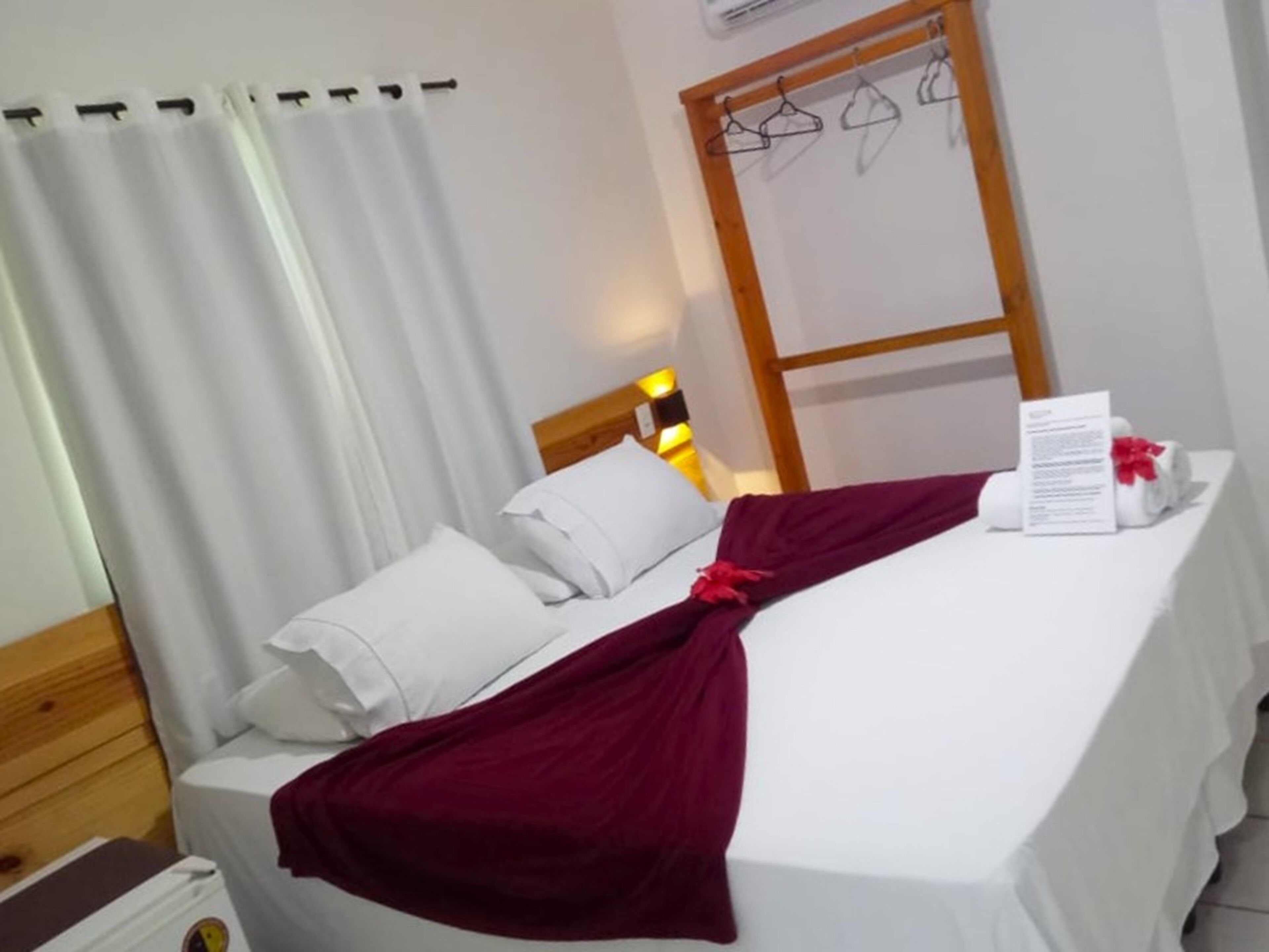 A neatly made bed with white linens, a red throw blanket, and a towel with a red flower. There is a wooden headboard, a white wall, and a window with white curtains.