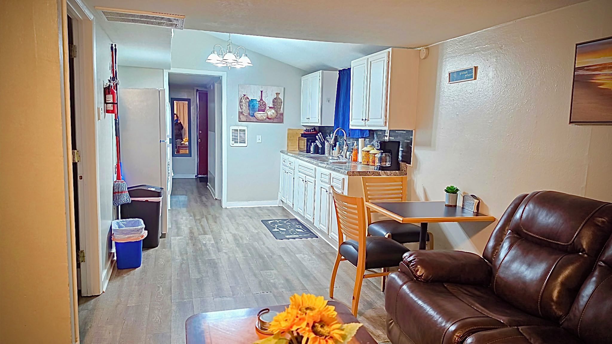 A living room with a brown leather sofa, a dining table with a chair, and a kitchen area with cabinets and a countertop.