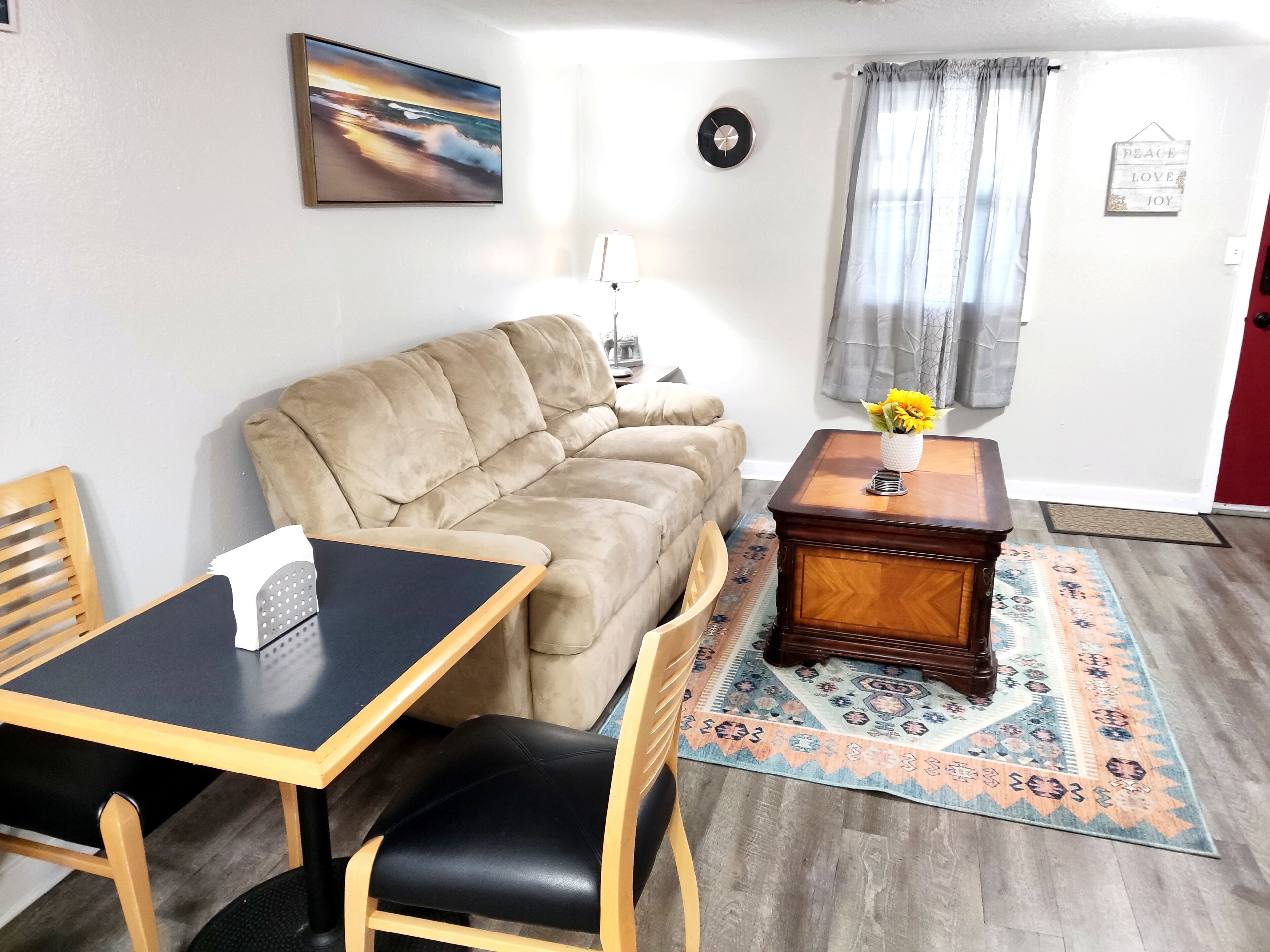 Ein Wohnzimmer mit einem beigen Sofa, einem Couchtisch aus Holz mit einer Vase voller Sonnenblumen, einem schwarzen Tisch mit einer Taschentuchbox und einem gemusterten Teppich.