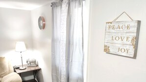 Ein Zimmer mit einem beige gefärbten Sessel, einem kleinen Tisch mit einer Lampe, einer dekorativen Elefantfigur und einem Schild an der Wand mit der Aufschrift 'PEACE LOVE JOY'.
