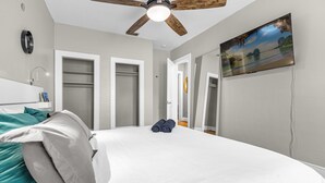 A bedroom with a large bed, a ceiling fan, a TV on the wall, and a view of the outdoors through a window.