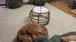 Een woonkamer met een groot schilderij, twee groene fauteuils, een ronde salontafel en uitzicht op een ondergaande zon door een raam.