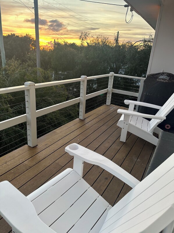 A wooden deck with white Adirondack chairs and a grill, overlooking a natural landscape with trees and a sunset.