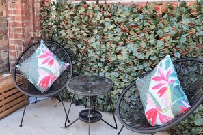 Two black wicker chairs with floral cushions, a small round table, and a dense green ivy wall.