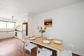 A modern kitchen with white cabinets, a wooden dining table set for four, and a vase with flowers on the wall. - Sun and Sand Albert Park (Albert Park)