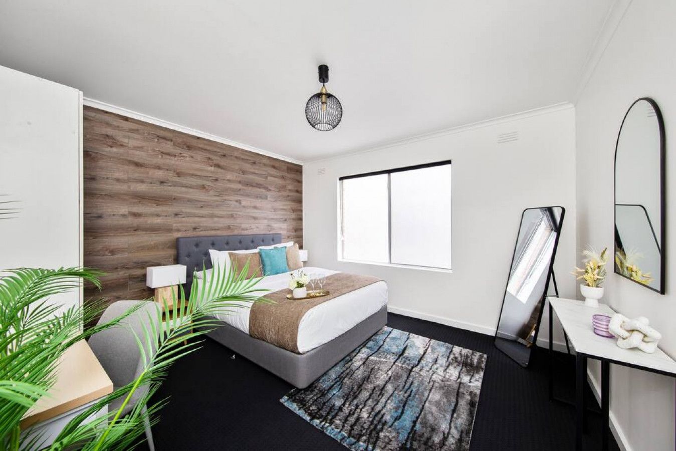 A modern bedroom with a wooden accent wall, a large bed, a bedside table with a mirror, and a view of the outdoors.