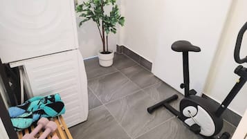 Ein kompakter Raum mit grauen Fliesen, einer weißen Tür, einem schwarzen Heimtrainer und einem kleinen Topfpflanzen.