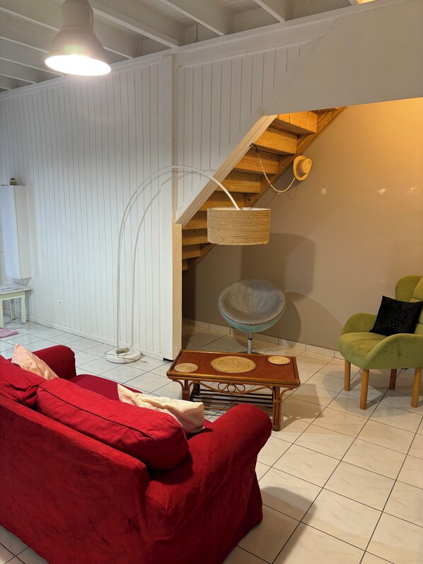 A room with a red sofa, a wooden coffee table, a green armchair, a woven basket, and a staircase with wooden railings.