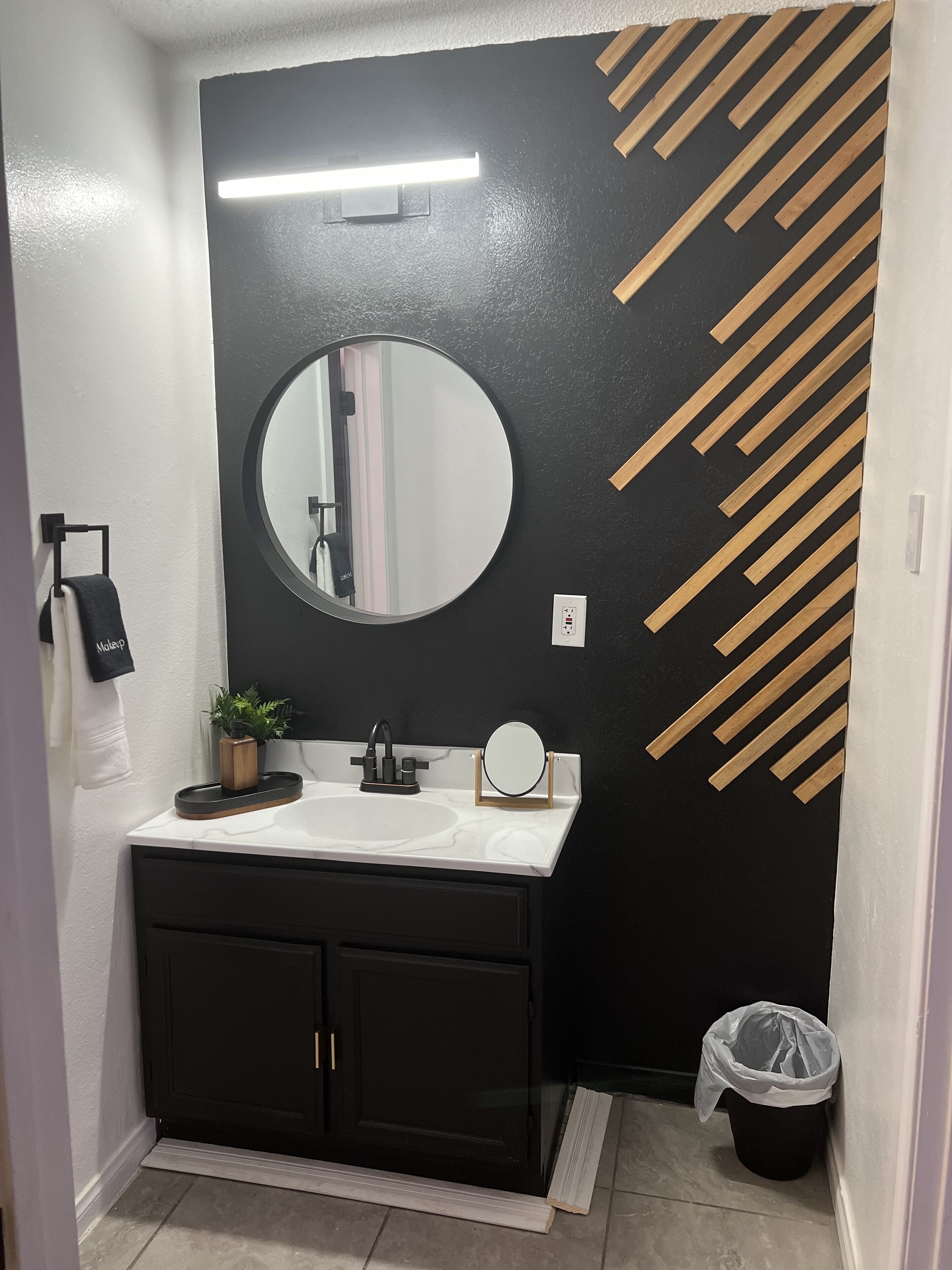 A bathroom with a black and white color scheme, a round mirror, a sink, a small plant, and a trash bin.