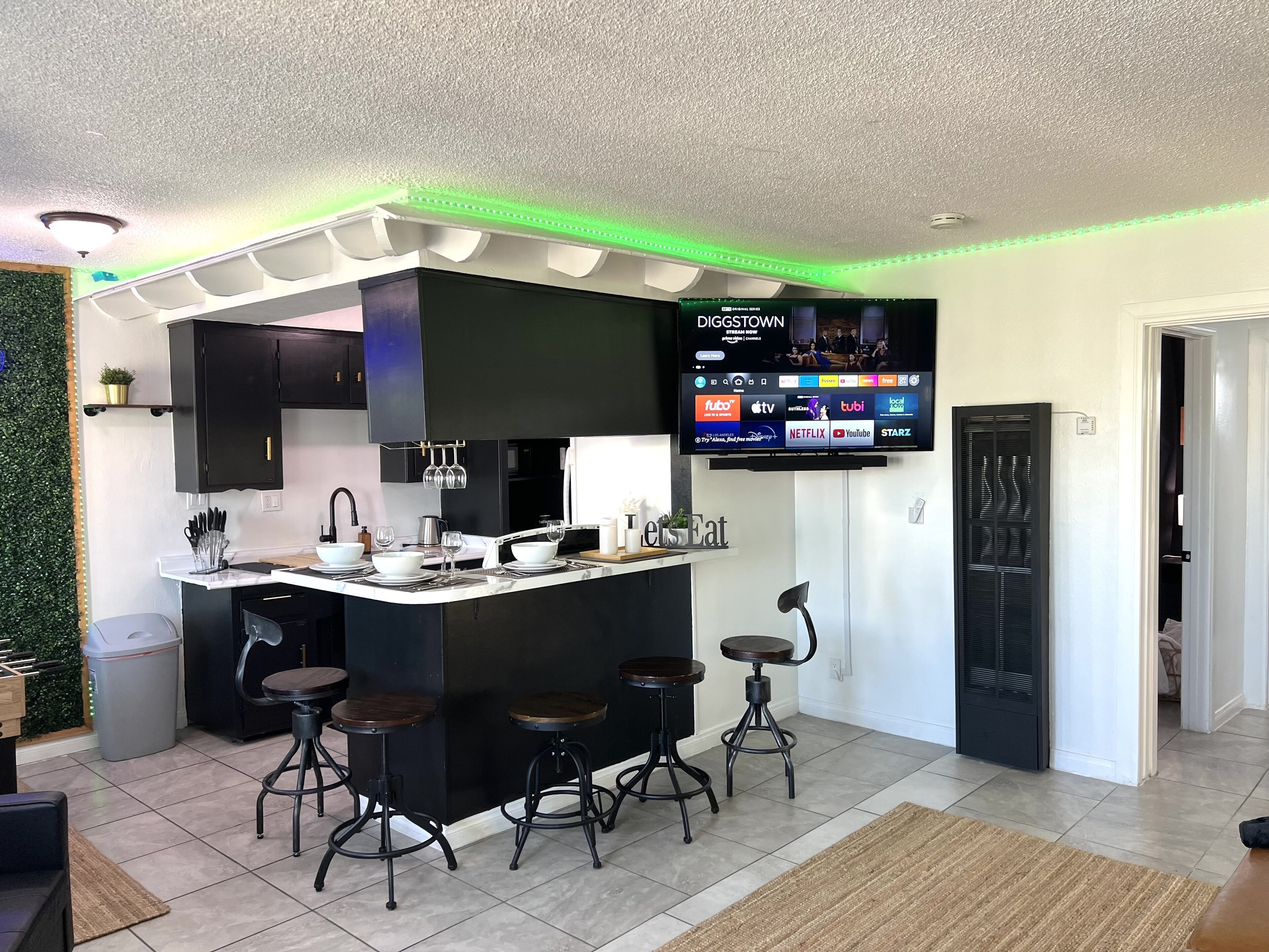 A modern kitchen with a central island, bar stools, a flat-screen TV displaying streaming services, and a green accent wall.