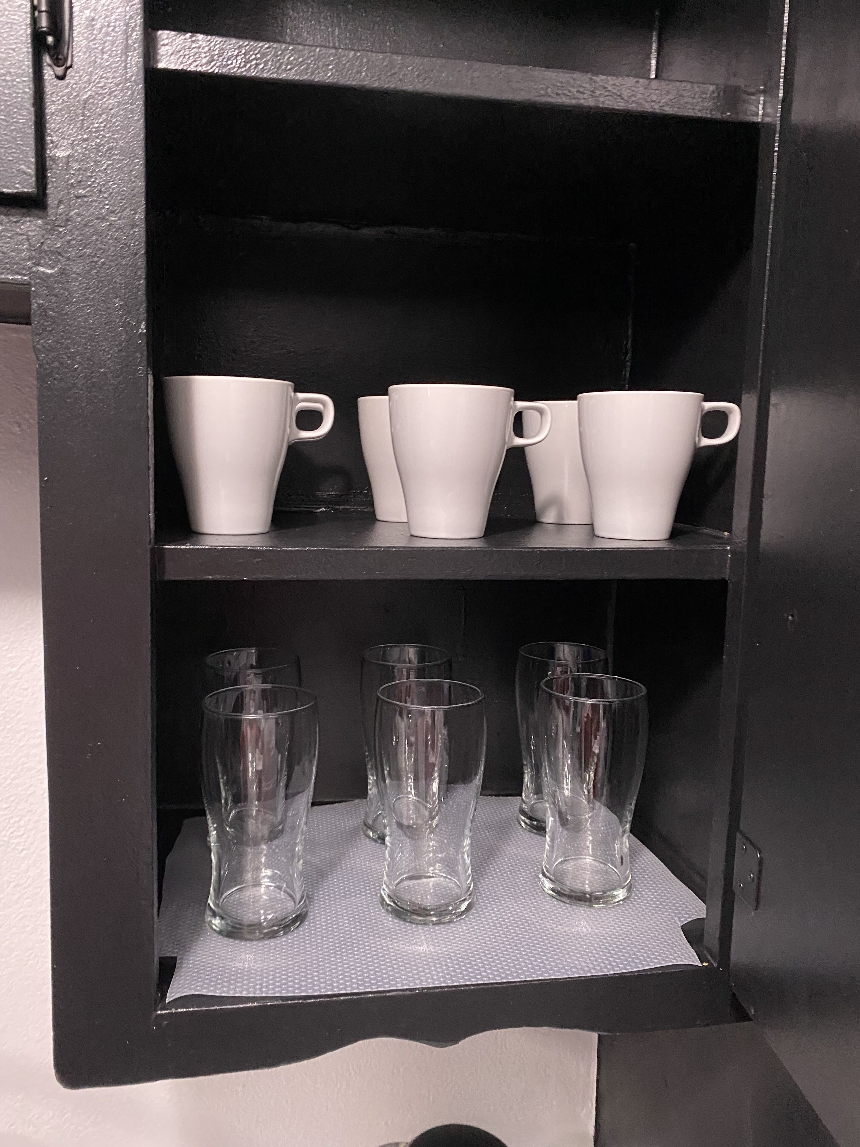 Un mueble negro con tres tazas blancas y tres vasos transparentes sobre estantes.