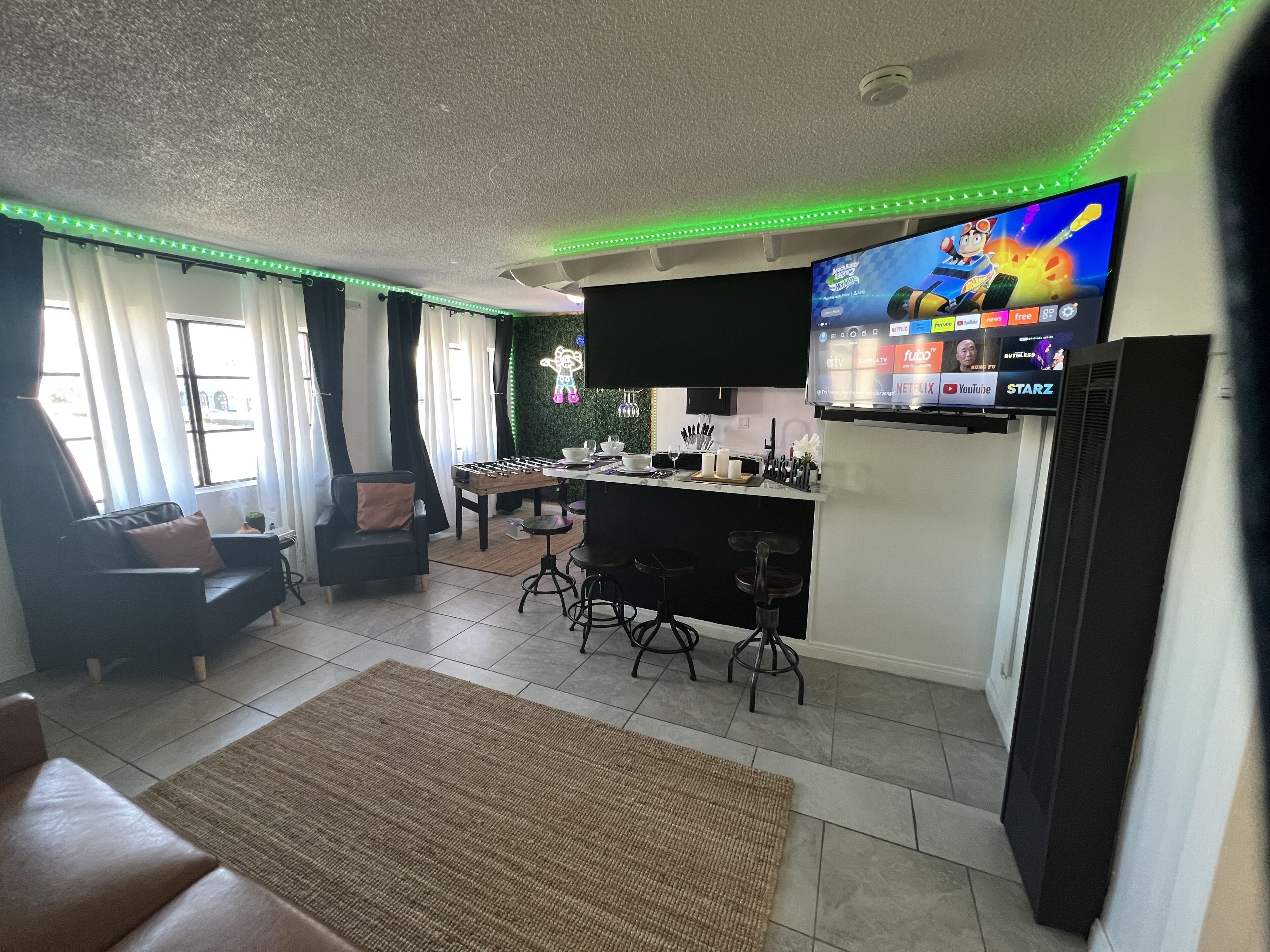 Ein modernes Wohnzimmer mit einer Couch, einem Couchtisch, einer Küchenzeile mit Barhocker und einem an der Wand montierten Fernseher, der verschiedene Sender anzeigt.