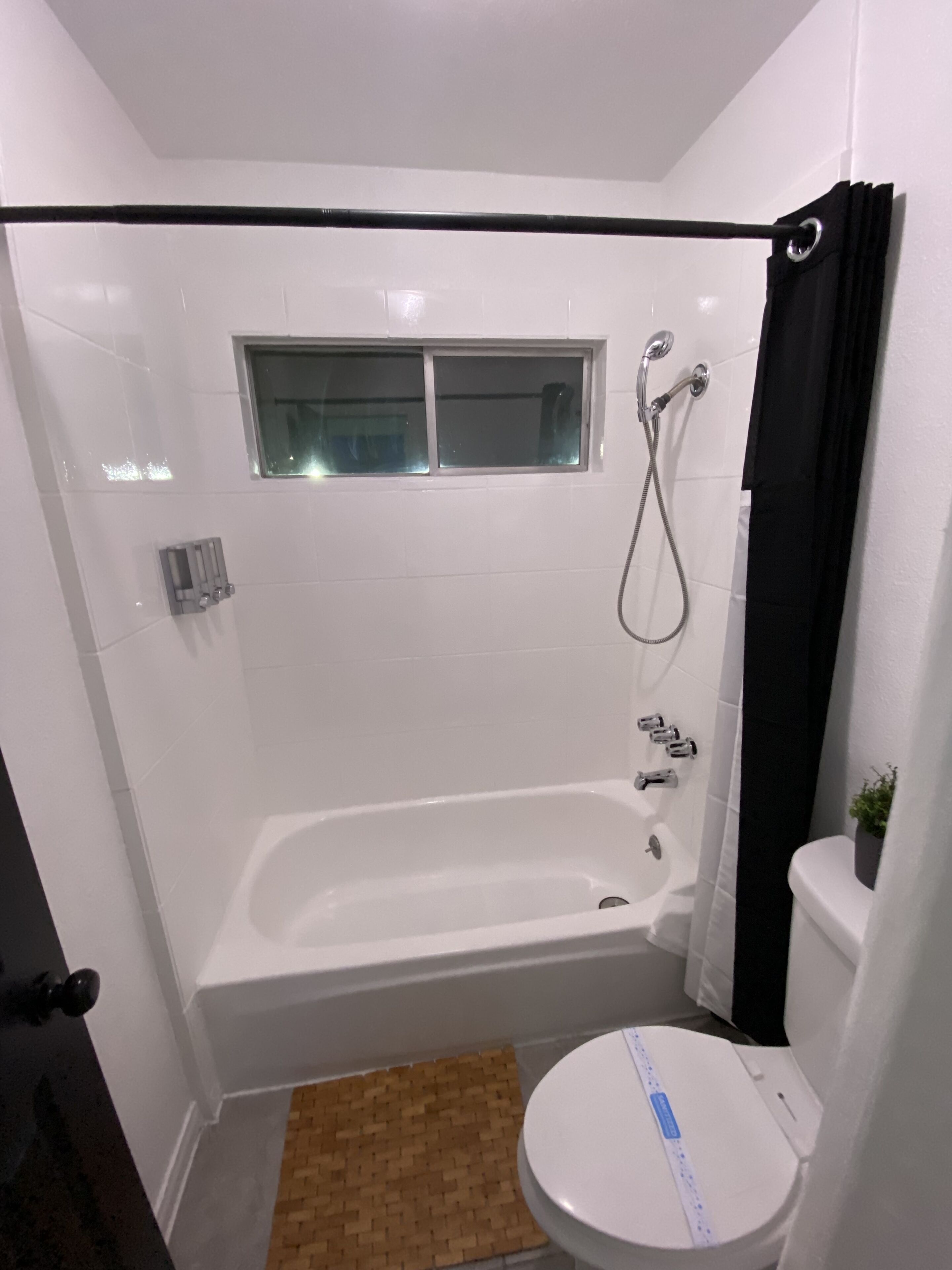 Een kleine, witte badkamer met een bad, een raam en een toilet.