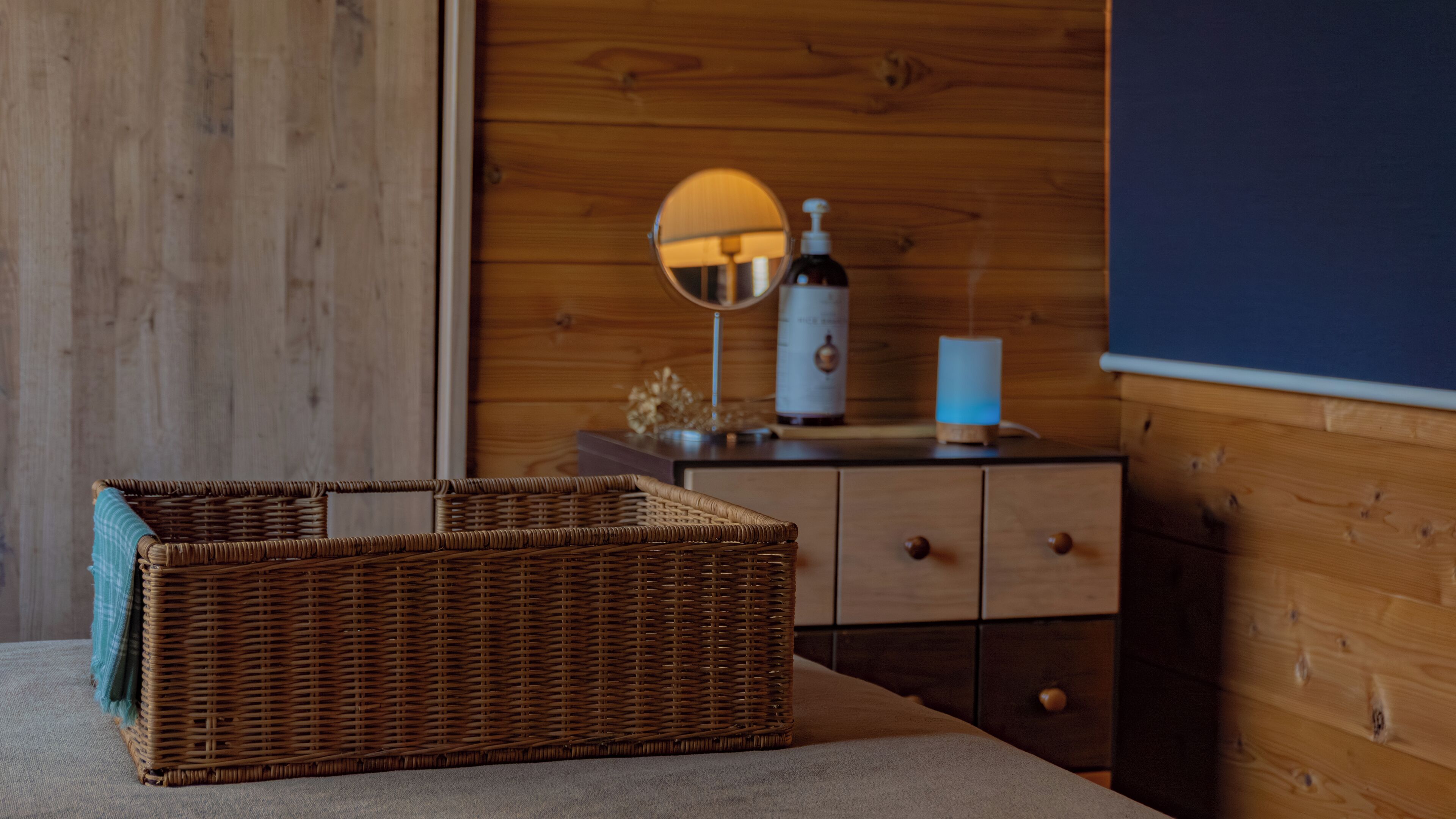 A wicker basket on a table, a mirror, a bottle, and a blue lamp against a wooden wall.