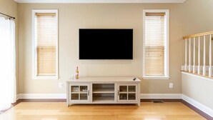 A room with a flat-screen TV mounted on the wall, a wooden floor, and a white console table with glass doors. - Charming 3BR & 2.5Ba nested in KOP w free parking, close to Conshy & Philly. (Bridgeport)
